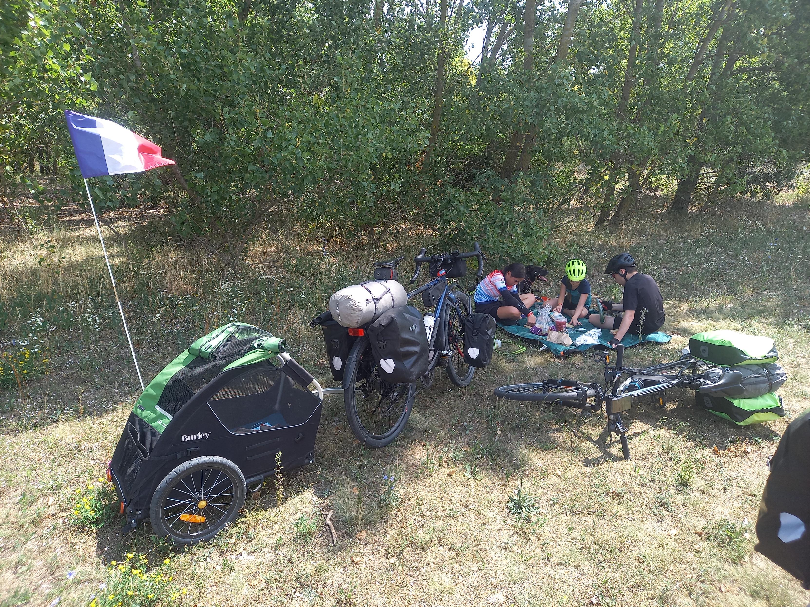 Un famille déjeune dans l'herbe. On voit des vélos chargés dont 1 avec la remorque du chien