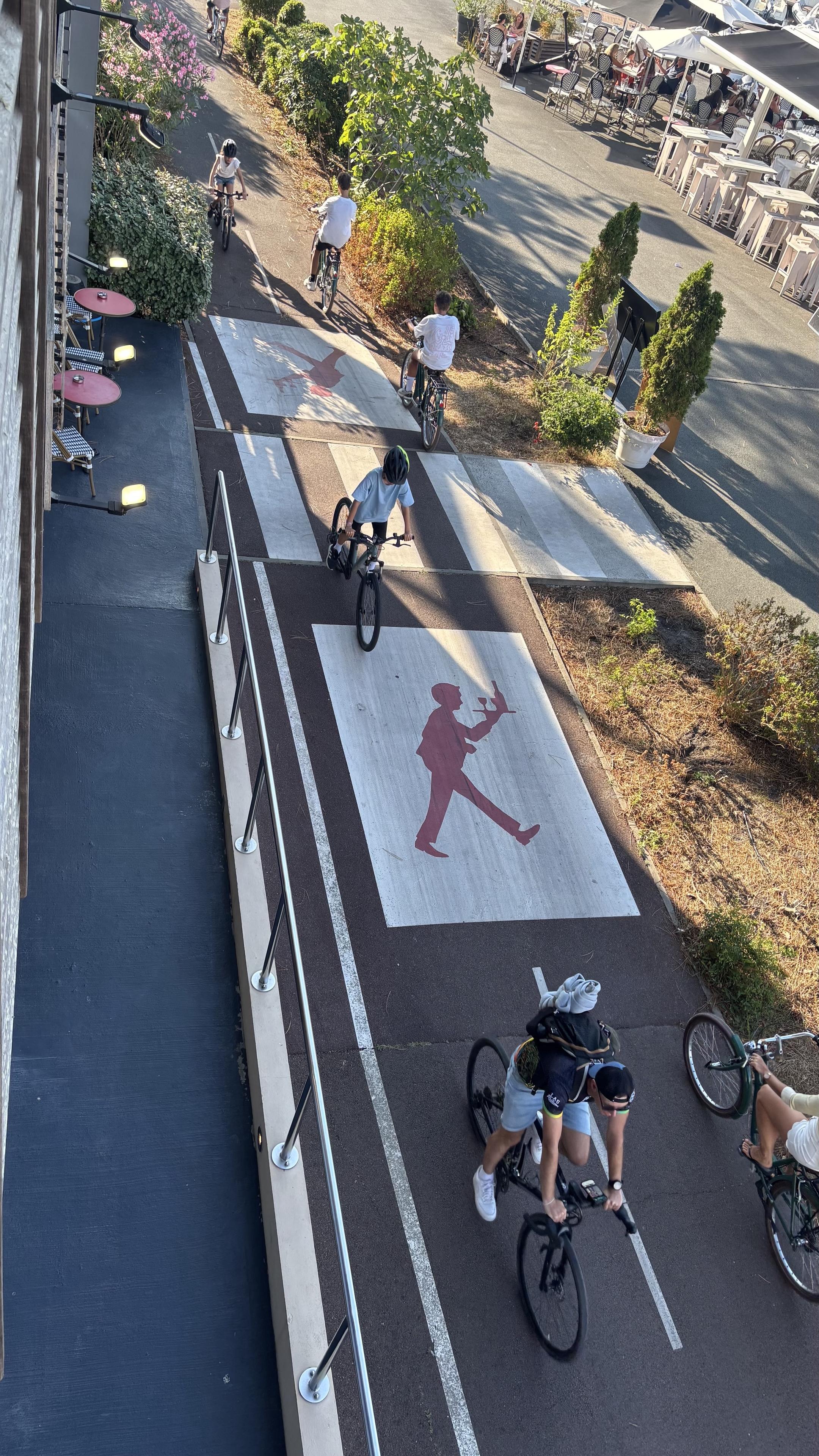 Sous un rayon de soleil des vélos passent en tous sens sur la piste cyclable qui est traversée par un passage piéton. Avant celui-ci, des deux côtés des bandes blanches un énorme pictogramme rouge sur fond blanc barré toute la largeur de la piste, il avertit de la présence d’un restaurant sous la forme d’un serveur stylisé qui traverse avec un plateau chargé posé en équilibre sur sa main.