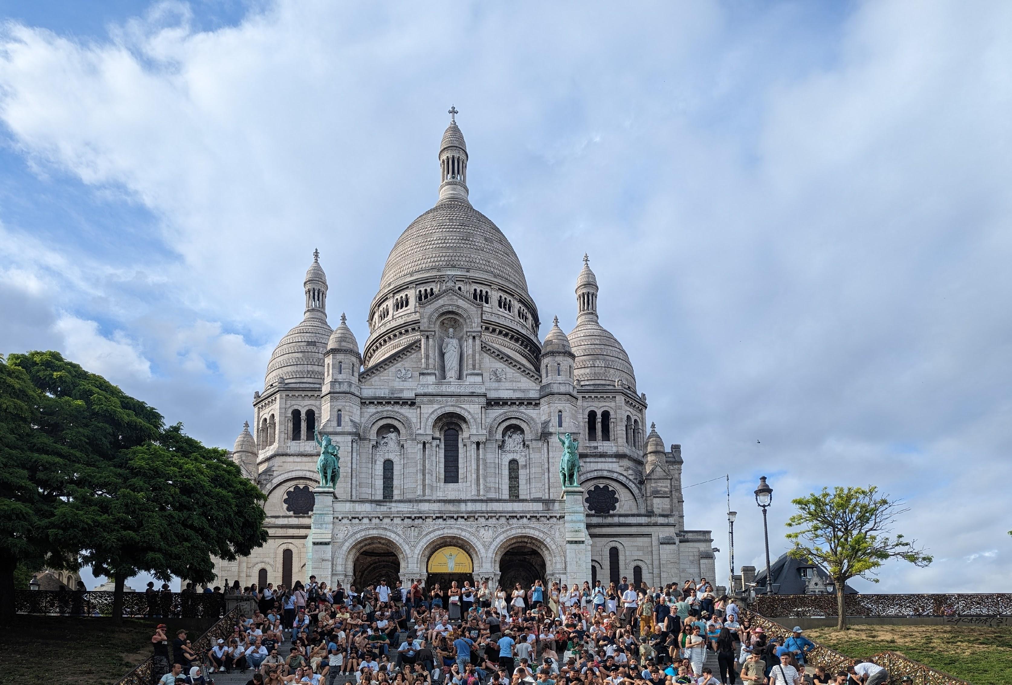 wide view of the Sacré-Cœur white marble basilica on top of the Montmartre hill in Paris. A huge central dome, two side smaller domes. A huge crowd on the stairs leading to the basilica.